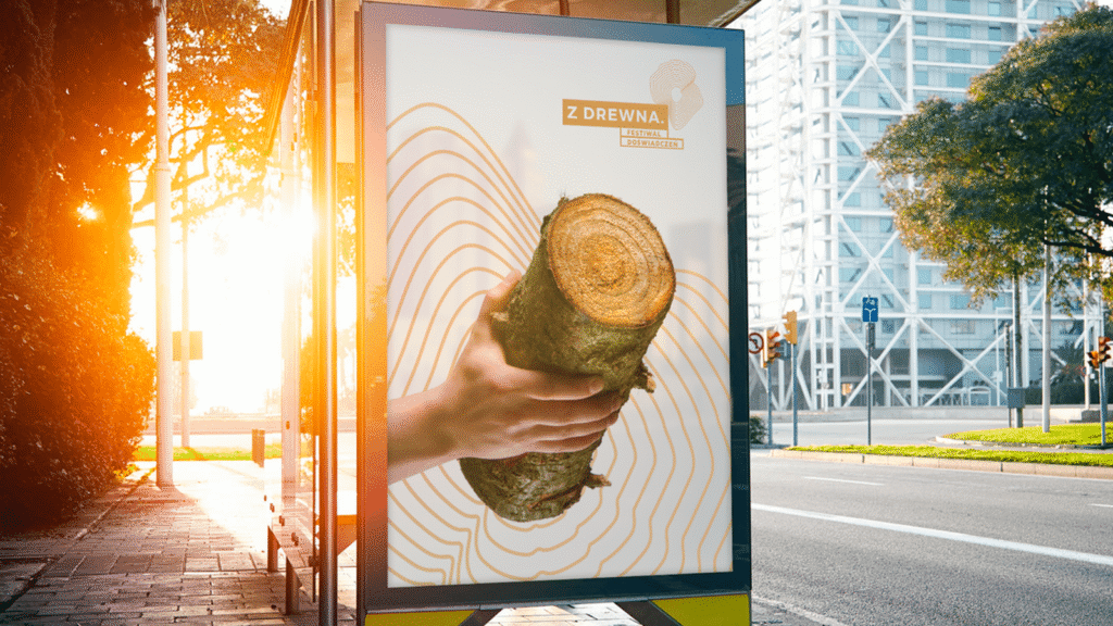 Plakat festiwalu z motywem drewna na przystanku autobusowym, oświetlony zachodzącym słońcem.